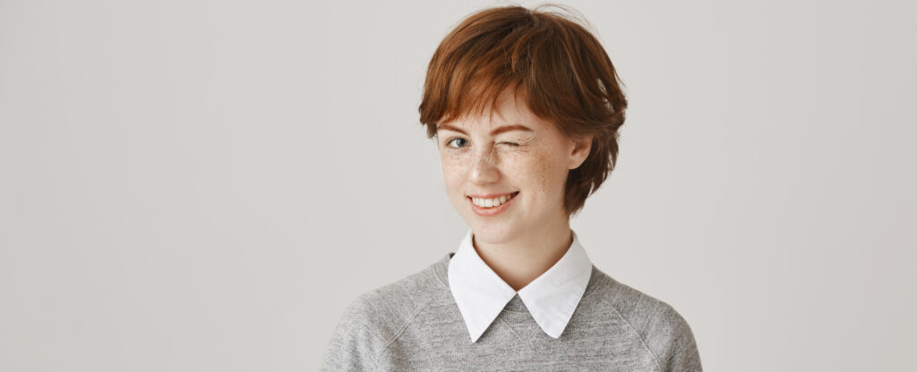 Essential Maintenance for Short Hairstyles if you know what i mean. studio shot of positive attractive redhead woman with freckles winking as if being on same line with friend, agreeing with idea, standing over gray background